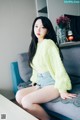 A woman sitting on a couch wearing a yellow sweater and denim shorts.